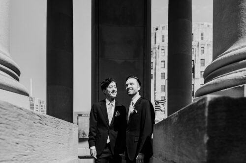 Two men in suits standing between stone pillars, smiling and looking upward.