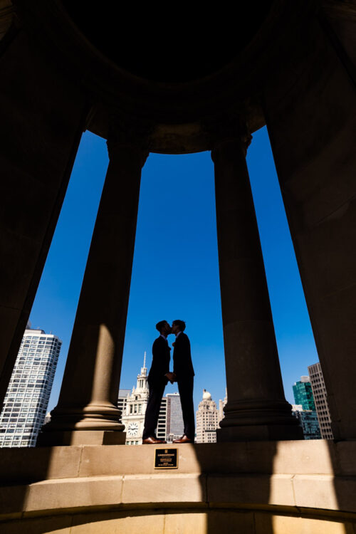 Silhouette of a couple kissing while standing between two large pillars against a bright blue sky.