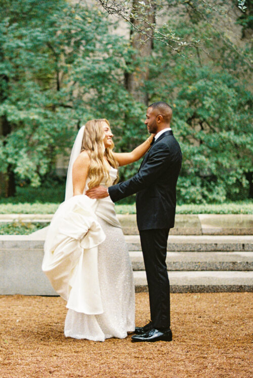 A bride in a white wedding gown and a groom in a black suit smiling at each other outdoors.