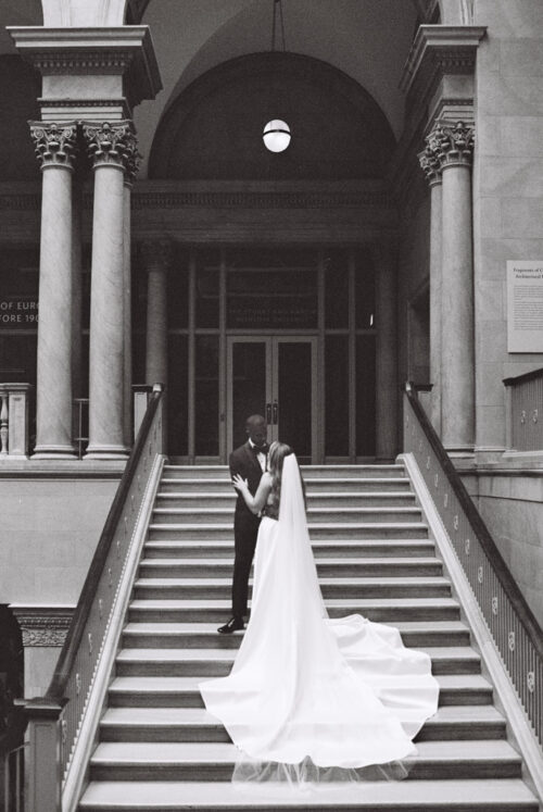 A bride and groom standing on the stairs of a grand building entrance.