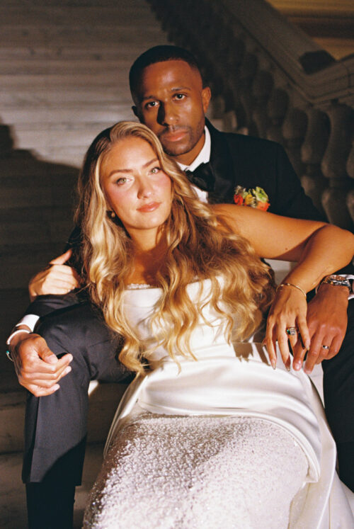 A bride in a white gown and a groom in a tuxedo sitting together on stone stairs.