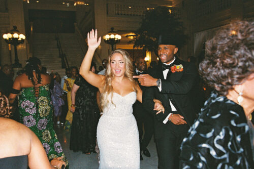 A bride and groom dancing at a reception, with the bride waving and the groom pointing.
