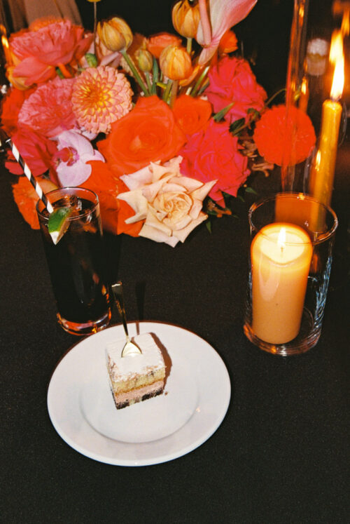 A slice of layer cake on a plate, a cocktail with a lime wedge, and lit pillar candles next to a floral centerpiece.